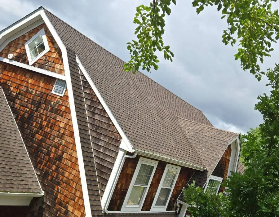 Upward angle view of a steep-pitch architectural shingle roof replacement on a natural cedar shake home with white fascia and dormer trim