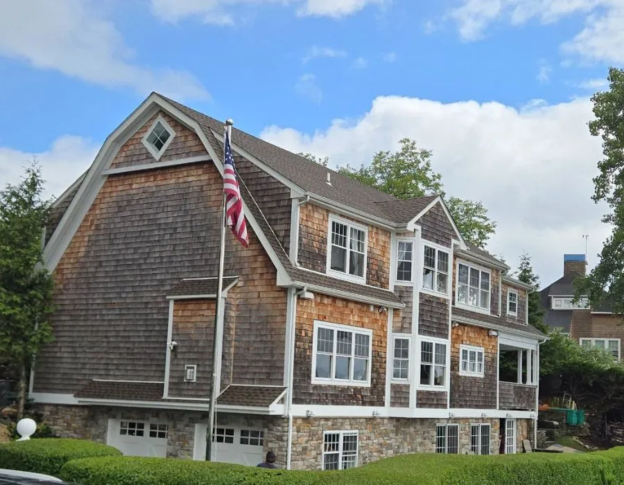 Side view of a multi-story natural cedar shingle house featuring a gambrel roofline, stone veneer foundation, white-trimmed windows, and an American flag flying on a flagpole out front