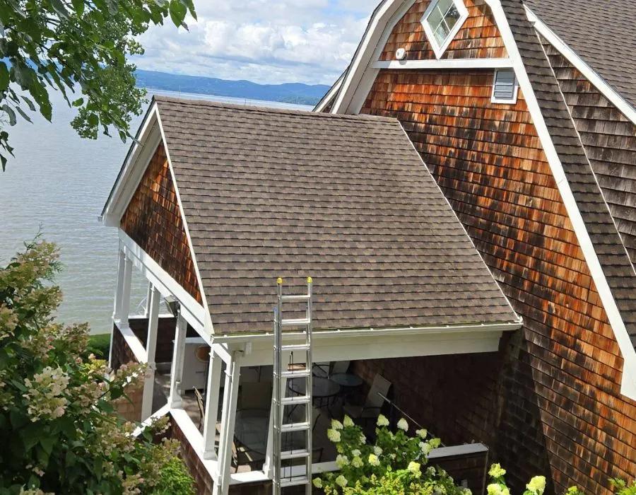 Elevated view of a brown architectural shingle roof covering a porch with a ladder leaning against it, set against a scenic waterfront backdrop and natural cedar shake siding